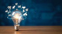 Glowing light bulb with business icons floating above on dark blue background representing innovation, creativity and entrepreneurial ideas for corporate success.