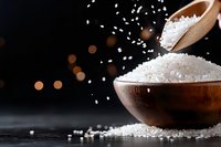 Sea salt crystals falling from wooden spoon into rustic bowl on dark background with bokeh lights and scattered grains on surface. Food photography.