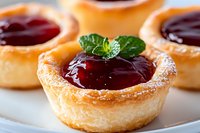 Mini pastry tarts filled with bright red berry jam and garnished with fresh mint leaves, dusted with powdered sugar on white plate, macro food photography.