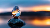 Crystal glass sphere on stone reflecting dramatic sunset sky and water surface with mirror reflection. Artistic nature photography with selective focus.