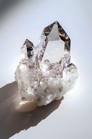Clear quartz crystal cluster with multiple points catching light and casting shadows, showcasing natural geometric formations and prismatic reflections on gray background.