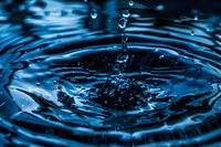 Macro shot of water drop splash creating concentric ripples on dark blue water surface with floating droplets and dynamic splash formation.