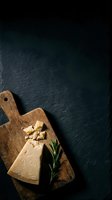 Wedge of aged cheese with crumbles on wooden cutting board and fresh rosemary sprig against dark slate surface for food styling and culinary content.