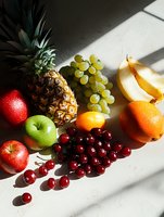 Fresh tropical and seasonal fruits arranged on white surface with dramatic natural lighting and shadows, including pineapple, cherries, apples, oranges and grapes.