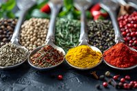 Colorful array of spices and herbs in metal measuring spoons, including turmeric, paprika, peppercorns, and dried herbs arranged on dark background.