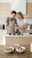 Happy young Caucasian couple cooking healthy smoothie together in modern kitchen wearing matching aprons while preparing nutritious breakfast meal.
