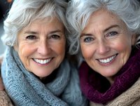 Senior Caucasian women with silver hair wearing winter scarves smile warmly at camera, showing genuine friendship and joy. Natural lighting highlights their confident expressions.