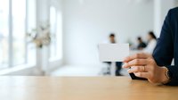 Man in dark business suit holding blank white business card at wooden table with blurred colleagues in background, suitable for corporate identity and professional networking concepts.