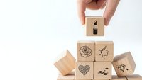 Hand placing wooden cube with lipstick icon above beauty and cosmetics symbol blocks on white background for branding and marketing concepts.