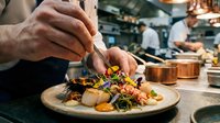 Chef plating gourmet dish with microgreens and sauce in professional kitchen restaurant setting.
