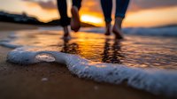 Romantic couple walking barefoot on beach at sunset with gentle wave foam in foreground, silhouetted legs against golden orange sky reflected in wet sand.