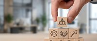 Hand placing wooden block with people icon on top of pyramid stack with business symbols including gear, handshake, and arrow for leadership and teamwork concepts.
