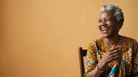 Senior woman with gray hair and dark skin tone, wearing a colorful patterned top, laughing joyfully against a warm yellow background. Suitable for healthcare, wellness, and lifestyle content.