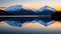 Snow-capped mountain peaks reflect perfectly in calm lake waters during golden sunset, creating mirror symmetry with dramatic winter landscape and peaceful atmosphere.