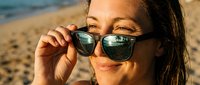 Young woman with dark hair and olive complexion adjusting black-framed sunglasses on sandy beach with ocean backdrop, summer vacation and leisure lifestyle concept.