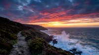 Dramatic coastal cliffs with crashing waves during vibrant sunset, golden and orange sky reflecting on ocean water, rugged rocky shoreline landscape for travel and nature backgrounds.