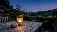 Glowing vintage lantern on rustic wooden table at twilight, with stone wall and rolling hills under a starry night sky.