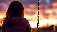 Young woman in purple coat looking at sunset, silhouetted against golden sky with chain swing in foreground, creating nostalgic mood.
