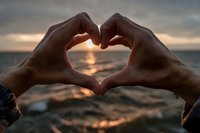 Hands forming heart shape silhouette against golden sunset over ocean waves, romantic gesture expressing love and connection during peaceful evening moment.