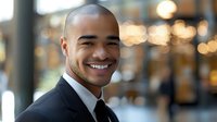 African American businessman with confident smile wearing black suit and tie against blurred office background with bokeh lights, expressing success and leadership.