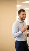 Young Caucasian male professional in light blue shirt and dark trousers gesturing while speaking in modern office corridor with warm lighting.
