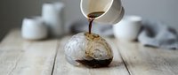 Pouring hot coffee into a cracked eggshell on wooden surface with blurred kitchen items in background, suitable for creative advertising and beverage concepts.