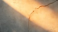 Dramatic diagonal sunbeam illuminates textured concrete wall with deep crack, creating contrast between light and shadow in minimalist architectural detail.