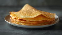 Homemade thin golden crepes stacked on white plate against dark background, close up view of traditional French pancakes with delicate lacy texture.