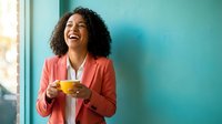 Happy African American businesswoman laughing while holding yellow coffee cup in modern office with turquoise wall and natural window light.