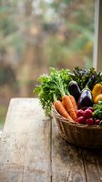 Wicker basket filled with fresh organic vegetables including carrots, eggplant, radishes and leafy greens on rustic wooden table by window with blurred garden background.