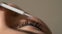 Close-up of woman applying white eyelash serum with applicator wand to long dark lashes for beauty skincare routine and cosmetic enhancement treatment.
