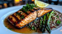 Grilled salmon fillet with caramelized glaze served alongside quinoa and fresh asparagus spears, garnished with parsley and lemon on white plate in restaurant setting.