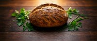 Freshly baked artisan sourdough bread loaf with crispy golden crust on rustic wooden table with fresh green herbs for bakery and cooking concepts.