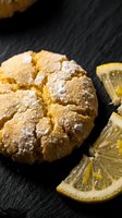 Fresh baked lemon crinkle cookies dusted with powdered sugar on dark slate surface with citrus slices for gourmet dessert and bakery concepts.