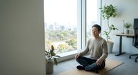 Young Asian woman practicing mindfulness meditation in modern apartment with city view, sitting cross-legged on yoga mat in peaceful morning light.