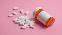 White prescription pills spilled from orange pharmacy bottle on pink background for healthcare and medication concepts.