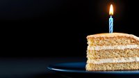Slice of layered vanilla cake with cream filling and lit blue birthday candle on dark background, dramatic lighting creates festive mood for celebration.