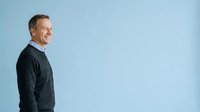 Middle-aged Caucasian male professional wearing dark sweater, smiling while looking over shoulder against blue background. Corporate portrait for business, leadership, and corporate communications.
