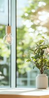 Minimalist home decor with wooden pendant light bulb and green plant in white ceramic vase against bright window background with soft bokeh effect.