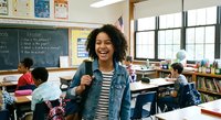 Young African American female teacher smiling in elementary classroom with students working at desks, educational environment with chalkboard and windows.