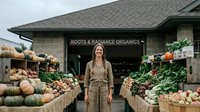 Young woman farmer standing proudly in front of organic produce market with fresh vegetables and pumpkins displayed on wooden stands at local farm store.