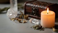 Mystical witch apothecary setup with burning candle, wooden chest, glass bottles and dried herbs on dark surface for magical rituals and spellcasting.