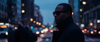 Man with dark skin tone wearing sunglasses and black jacket stands in urban street at dusk with blurred city lights and buildings in background, suitable for lifestyle, fashion, and urban themes.