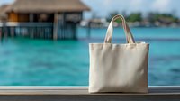 Beige canvas tote bag on wooden deck overlooking tropical turquoise lagoon with overwater bungalows for vacation travel and beach lifestyle mockup.