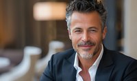 Confident mature businessman with salt and pepper hair and stylish beard wearing black blazer smiles warmly in modern office setting. Professional corporate portrait.