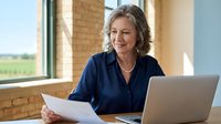 Mature Caucasian businesswoman smiling while reviewing documents at laptop in modern office with brick walls and large windows.