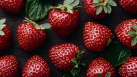 Fresh ripe strawberries with green leaves scattered on dark wooden background, macro shot showing detailed texture and natural pattern of red berries.