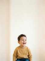 Young boy, approximately 3-4 years old, mixed ethnicity, wearing mustard yellow sweatshirt and blue jeans, laughing with genuine joy against neutral beige background. Studio portrait.