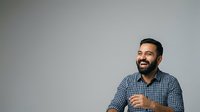 Male professional with dark hair and beard, wearing blue checkered shirt, smiling and gesturing while speaking against neutral gray background for corporate communications.