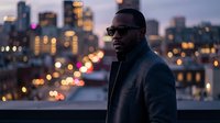 African American male wearing dark sunglasses and navy jacket standing on rooftop at dusk with illuminated cityscape behind him, suitable for lifestyle, fashion, and urban editorial use.